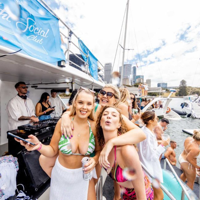 A group of people enjoying a private yacht charter in Sydney Harbour. Several women in swimsuits pose for a photo while a man DJs. Other sailboats and buildings are visible in the background, making it an ideal scene for corporate boat events Sydney.