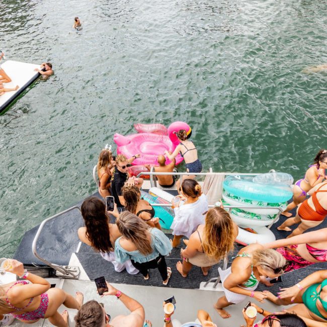 A group of people in swimwear are gathered on a private yacht charter in Sydney Harbour, some holding drinks. Several floatation devices, including a pink flamingo, float in the water near the boat. One person is swimming nearby, adding to the fun atmosphere of this perfect Catamaran party Sydney experience.