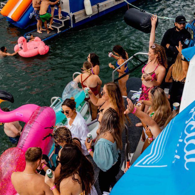 People are gathered on a boat and in the water, some on pool floats, relaxing and socializing. The boat has a banner with text and a water slide. Many individuals are holding drinks. It's the perfect scene for a Catamaran party Sydney experience or DJ boat hire Sydney adventure.