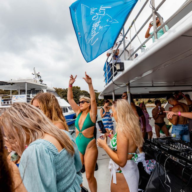 People enjoying a party on a catamaran, with one person wearing a green swimsuit dancing near the sound equipment. The backdrop includes other boats and a cloudy sky. Perfect for corporate boat events Sydney or DJ boat hire Sydney.