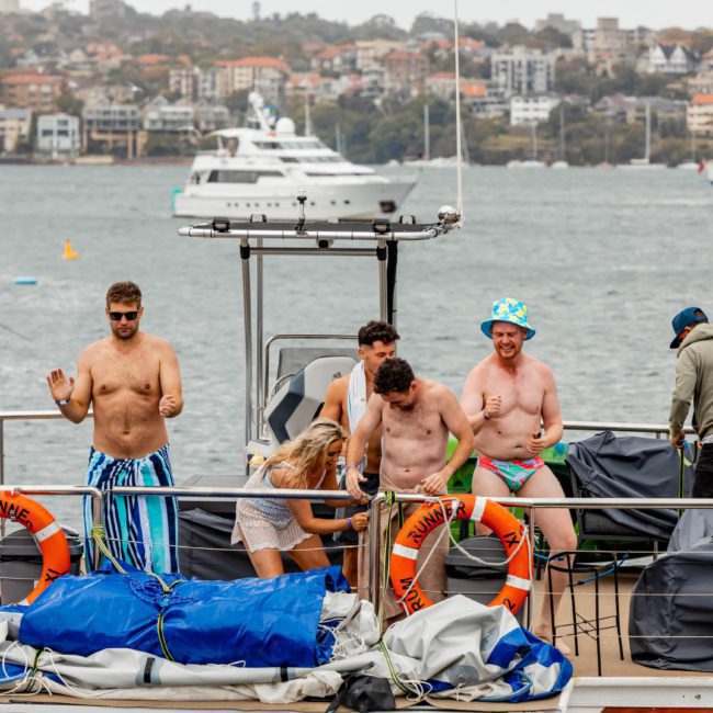 A group of people, some shirtless, stand and interact on the deck of a boat with lifebuoys. Other boats and the city shoreline are visible in the background, perfect for those considering a Sydney boat party hire.