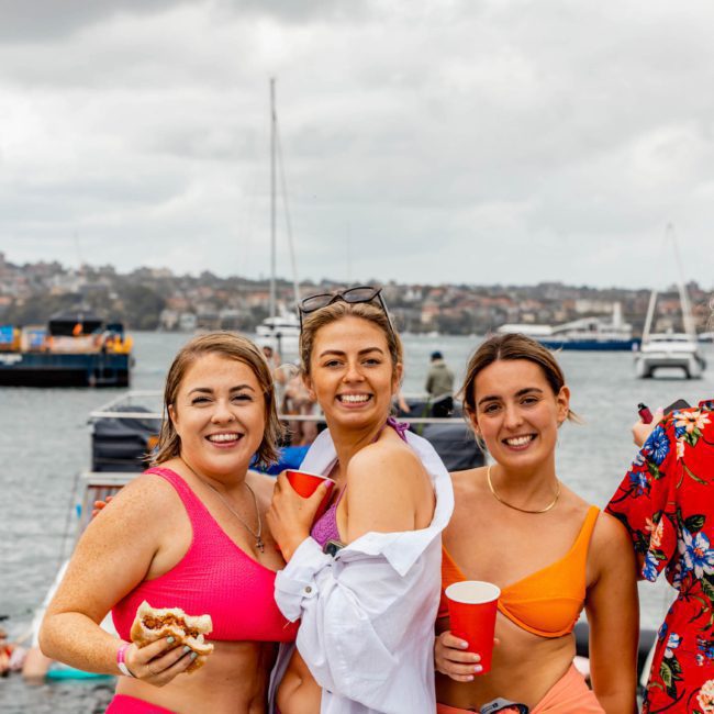 Three women in swimsuits and a partially buttoned shirt are smiling at the camera by the waterfront, holding food and drinks. Behind them, boats are anchored in the water under a cloudy sky, hinting at a festive Sydney boat party hire vibe.
