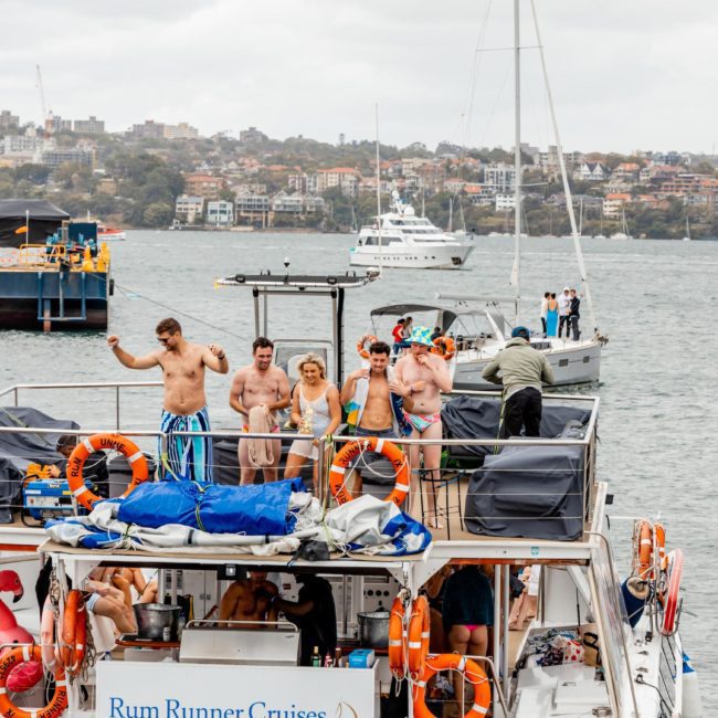 A group of people in swimsuits enjoying a catamaran party on the deck of a vessel named "Rum Runner Cruises" with more boats and a cityscape in the background.