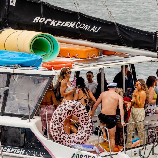 A group of people in swimwear are gathered on a docked sailboat, socializing. The boat is equipped with various colorful flotation devices and displays the brand "ROCKFISH.COM.AU" on the sails, perfect for a Sydney boat party hire.