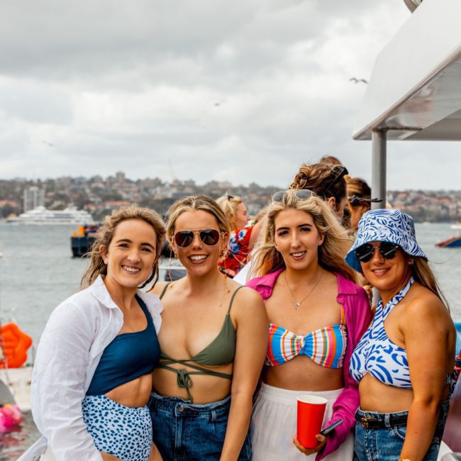 Four women wearing swimwear and sunglasses stand on a boat, smiling toward the camera. The background features a cloudy sky, water, boats, and a distant shoreline. Perfect for capturing memories at a private yacht charter in Sydney Harbour.