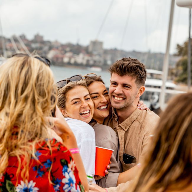 Three people standing close together, smiling and hugging, by a waterfront with boats in the background. A person in the foreground wears a red floral shirt. It's a perfect day for a private yacht charter on Sydney Harbour.