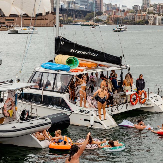 A group of people are enjoying a party on a luxury yacht hire Sydney, with floating devices on the water and the stunning Sydney Opera House and city skyline in the background.