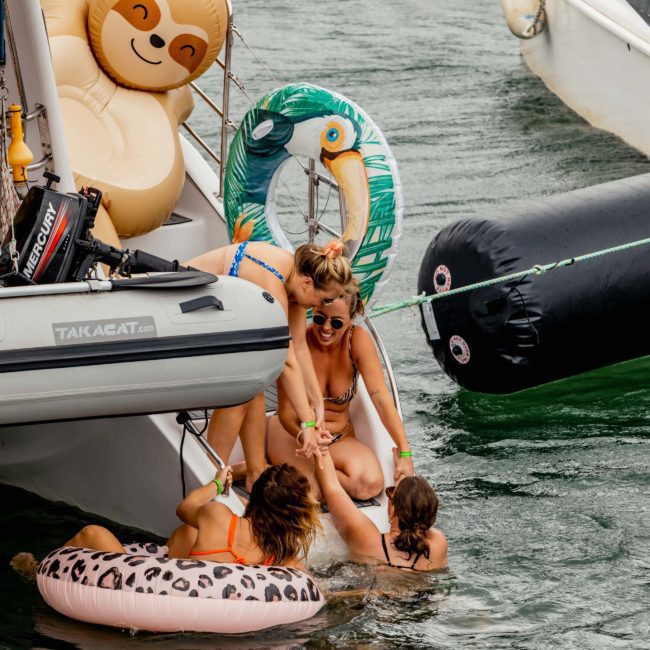People enjoying a day on the water with inflatables; one person is climbing a boat ladder while others assist. A sloth and toucan float are visible on the boat and water, enhancing the fun atmosphere of this Catamaran party in Sydney.