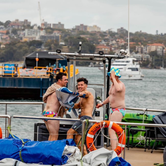 Three men in swimwear are on a docked boat, preparing for a swim. One man puts on a swim cap, while another is adjusting his swimwear. Various equipment and life preservers are visible on the boat, suggesting they're ready for an unforgettable day with DJ boat hire Sydney or catamaran party Sydney services.