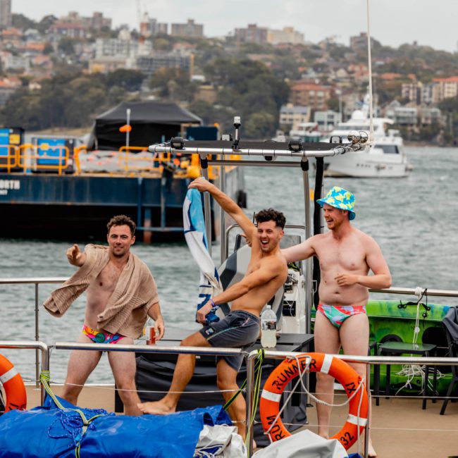 Three men in brightly colored swimwear and towels pose playfully on a boat deck near a city waterfront with various ships and buildings in the background, perfect for an unforgettable Sydney boat party hire.