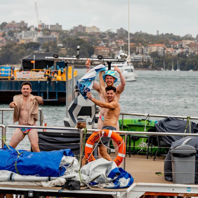 Four men on a boat by the shore. Three are shirtless, one in a bathrobe. Two are holding up a bicycle. The background shows water, buildings, and another boat—perfect for considering a luxury yacht hire Sydney experience.