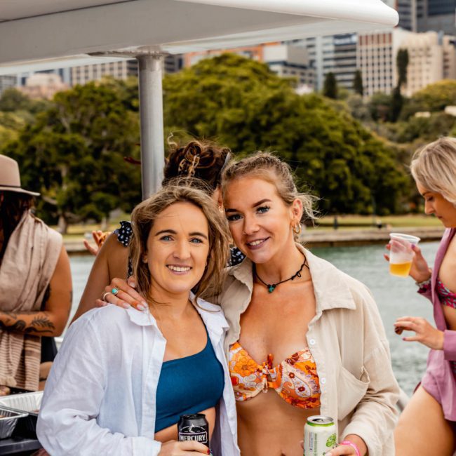 Two women are posing for a photo on a boat. The woman on the left is wearing a blue dress shirt, while the woman on the right sports a bikini top with an open shirt. Others in the background are enjoying the catamaran party.