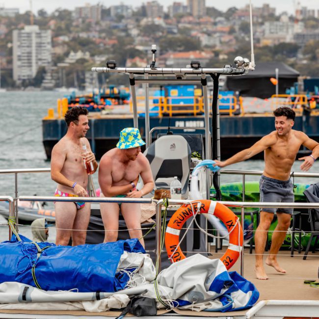 Three men on a catamaran party in Sydney wearing swimwear, two holding canned drinks, with one man sporting a colorful bucket hat. The boat is docked at a marina with lifebuoys and tarps, and a cityscape in the background.