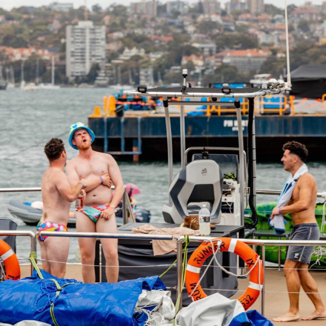 Three men are standing on a boat deck by the waterfront in Sydney Harbour. Two are in colorful swim trunks, one wearing a helmet. The third man, in a tank top and shorts, has a towel around his neck. Consider DJ boat hire or private yacht charters for an unforgettable experience on the water.