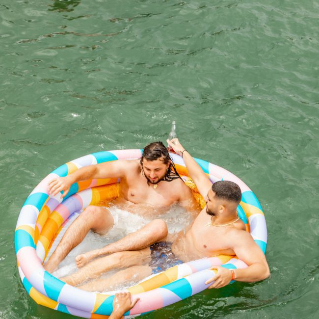 Three people are in a river, using inflatable devices to float. Two men are seated in a small inflatable pool, while another person is in an inner tube nearby. Another person is partially visible in the foreground, reminiscent of a relaxed moment from a DJ boat hire Sydney event.