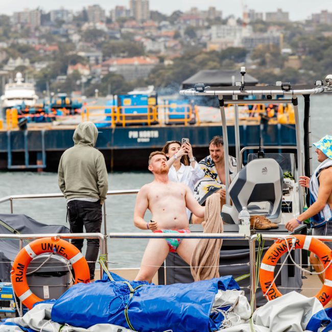 A group of people standing on a private yacht charter in Sydney Harbour with a cityscape and a barge in the background. One person in swimwear is striking a pose, while others are taking photos and attending to boat equipment.