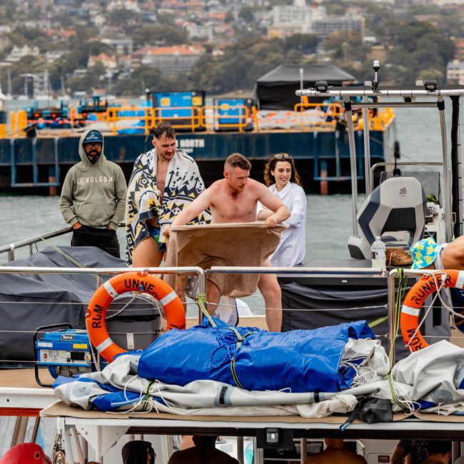 A group of people on a boat, with one person slipping over the edge. Various supplies and safety rings are visible on the deck, while the background shows water and docked vessels. Perfect setting for a catamaran party Sydney or DJ boat hire Sydney experience.
