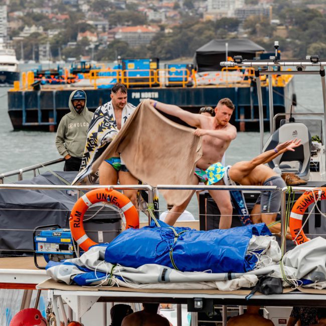 Three men in swim trunks are playing with a large towel on the deck of a boat, while another man observes. The background shows water, other boats, and city buildings. Life rings are visible on the railings, creating a perfect scene for luxury yacht hire Sydney.