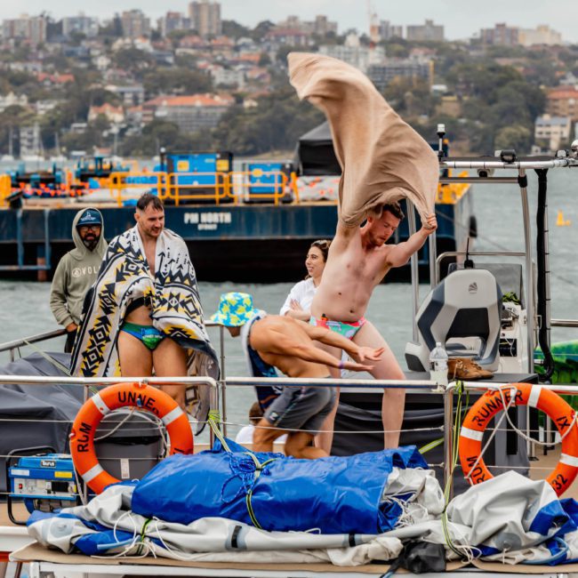 A group of people stand on a dock near the water. One person is leaping with a towel in the air, while others, some wrapped in towels, look on during a corporate boat event in Sydney. Various boats and buildings are visible in the background.
