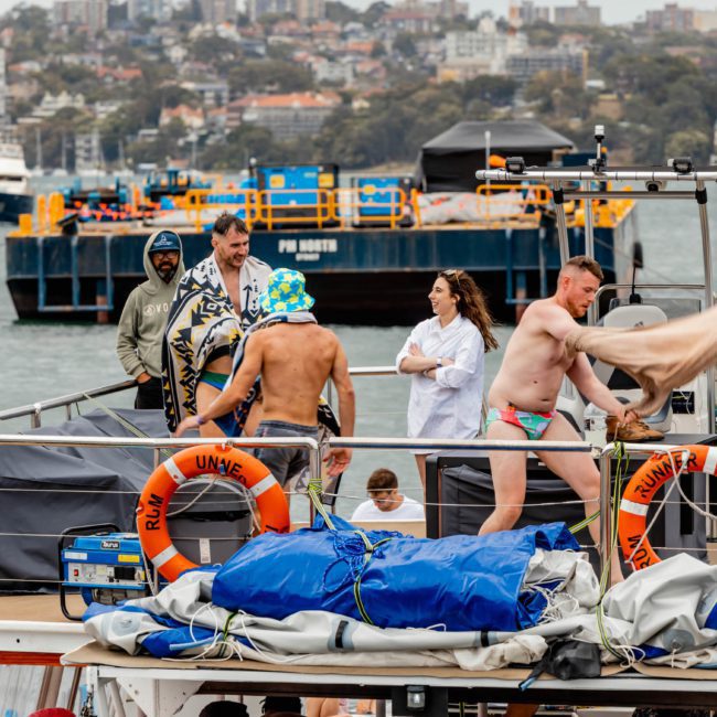 A group of people in swimwear, some with towels, stand and move around on the deck of a boat with lifebuoys labeled "MANTRA." The background shows water, boats, and buildings along the shore—perfect for a Sydney boat party hire.
