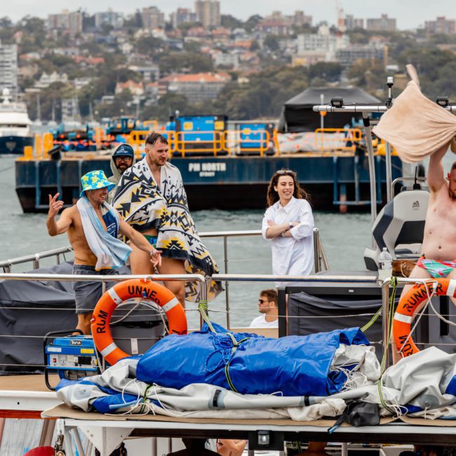 A group of people stand on a boat in a harbor, some wrapped in towels. Buildings and other boats are visible in the background, hinting at the vibrant atmosphere of a catamaran party Sydney.