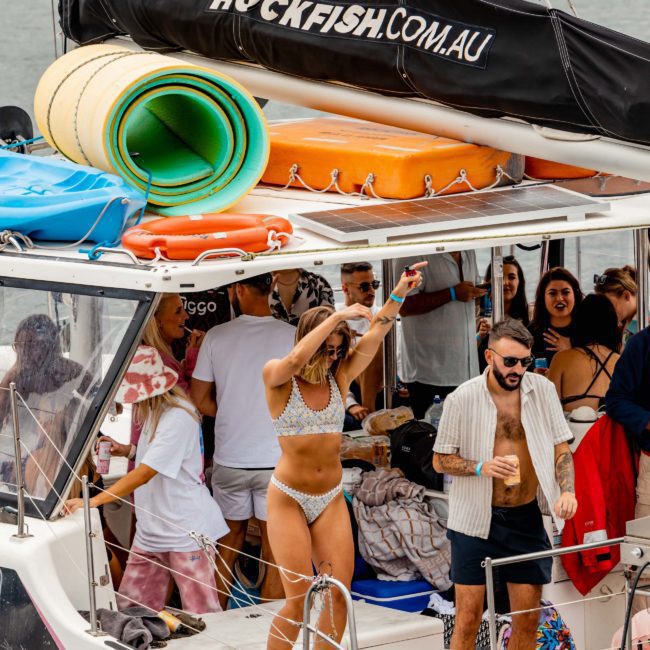 Group of people enjoying a Sydney boat party hire on Rockfish.com.au. A woman in a bikini is dancing, while others are socializing and holding drinks on this luxury yacht hire. The boat is docked on the sparkling waters of Sydney Harbour.