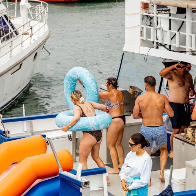 People on a boat engaged in various activities. Some are preparing to go down an orange slide, while others are talking and lounging. A person with a blue inflatable tube stands near the slide, all enjoying a luxury yacht hire in Sydney Harbour.