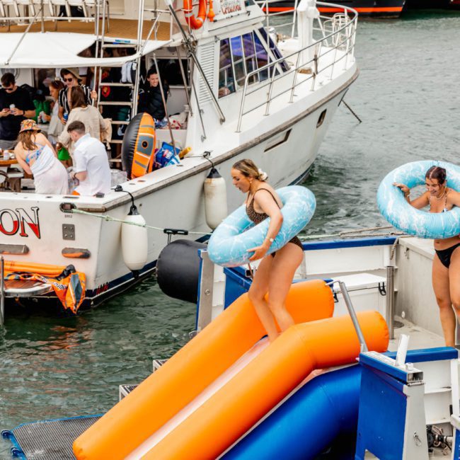 People enjoying a day on the water with boats and water slides. Most are in swimwear or casual attire, and one person is dressed as Santa. Two women are using the slides while others socialize on the boats, making it a perfect scene for a DJ boat hire Sydney event.