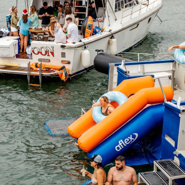 People enjoying a day on and around boats; some are dining on the boat named "MOON," while others use an orange inflatable slide and swim in the water. Ideal for corporate boat events Sydney, everyone is having a fantastic time under the sun.