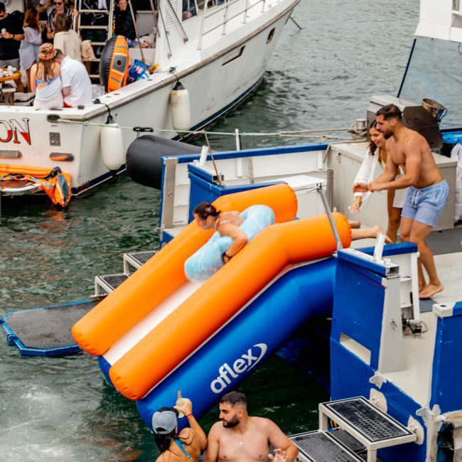People enjoy water activities on boats; some slide into the sea, while others chat and relax on a blue vessel. Nearby, more individuals gather for a Sydney boat party hire.