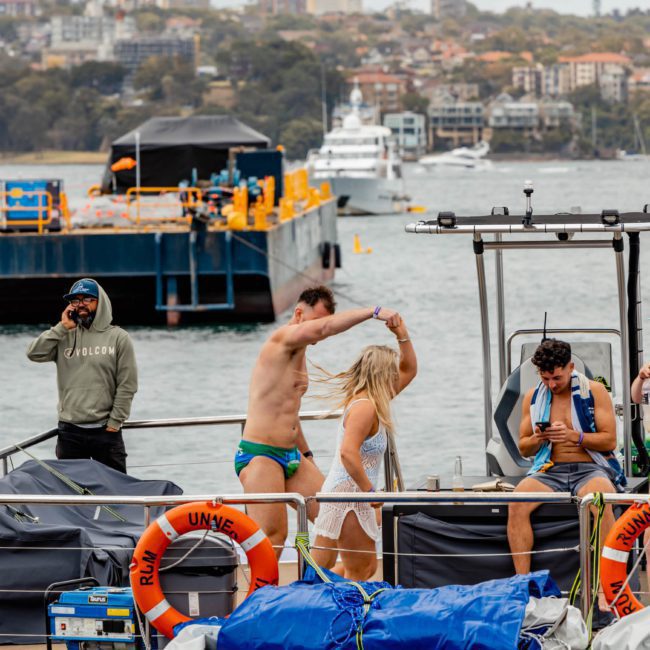 A group of people on a boat is having fun; two are dancing, one is on the phone, and another is watching. A person on the dock looks on. Residential buildings and another boat are seen in the background. Consider this scene for your next luxury yacht hire Sydney adventure!