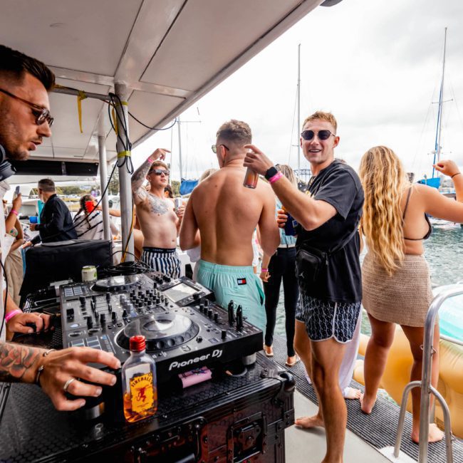 A group of people enjoying a Sydney boat party hire with a DJ playing music, individuals holding drinks, and luxury yachts visible in the background.