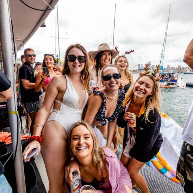 A cheerful group of people, mostly women, smiling and posing on a boat during a Sydney boat party hire. Some hold drinks with the picturesque marina visible in the background.