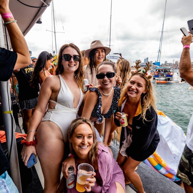 A group of people in swimwear and casual clothes enjoy drinks and smile on a boat by the water, with other boats and people visible in the background. Perfect for those seeking Sydney boat party hire or catamaran party Sydney experiences.