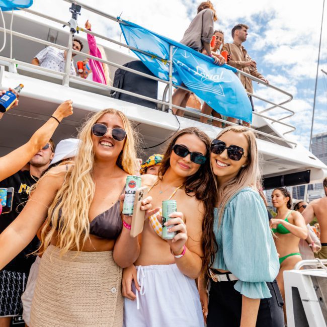 Three women wearing sunglasses are posing and holding drinks on a private yacht charter in Sydney Harbour, with other people enjoying the background.