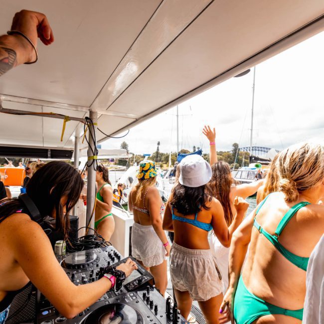 A group of people in swimwear enjoying a catamaran party in Sydney. A DJ is playing music as others dance and socialize. Another boat and a bridge are visible in the background, adding to the lively atmosphere.