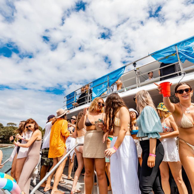 People socializing and holding drinks on a two-tiered catamaran under partly cloudy skies. Many are in swimwear, suggesting a festive or party atmosphere. A banner reads "Social Boat," indicative of a perfect Sydney boat party hire experience.