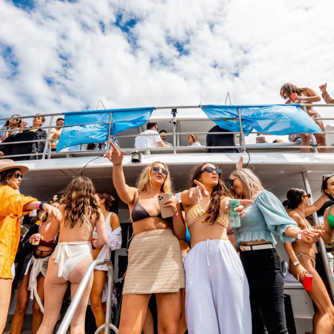 A group of people in swimsuits and summer clothes enjoying a festive atmosphere on a multi-deck boat with banners under a partly cloudy sky during a catamaran party in Sydney.