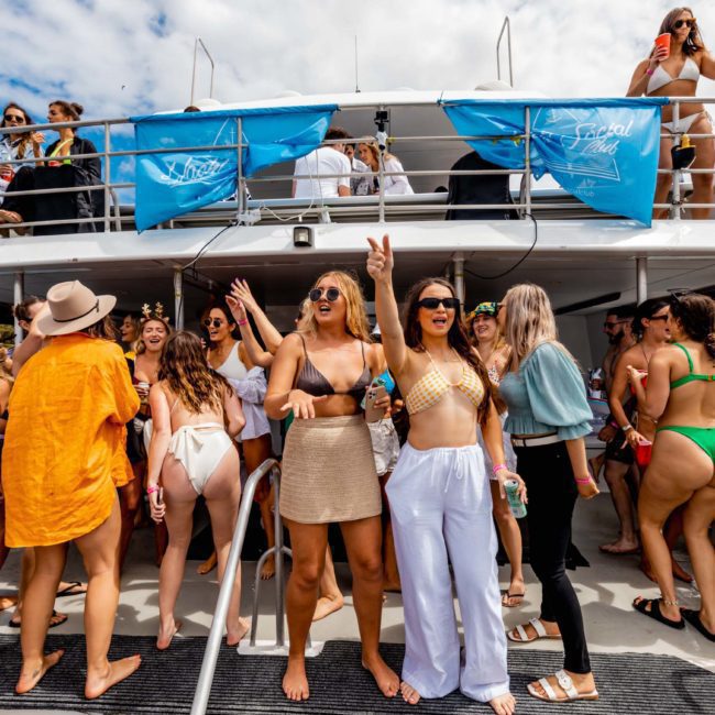 A group of people in swimsuits and summer outfits are gathered on a catamaran for a party, with some on the upper deck and others on the lower deck, enjoying the sunny weather.