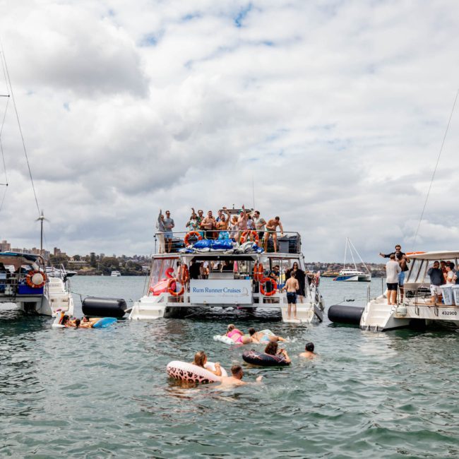 Three boats are anchored close together in a body of water, with people on board and in the water around them. Several individuals are floating on inflatables, enjoying a Sydney boat party hire experience under the cloudy sky.