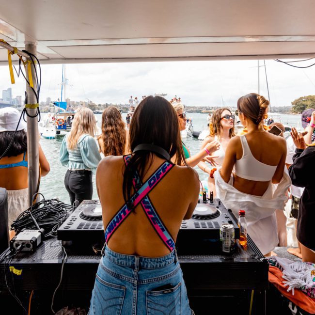 Experience a DJ performing on a luxury yacht with a crowd of people dancing and socializing under a covered area on a sunny day, ideal for corporate boat events in Sydney.