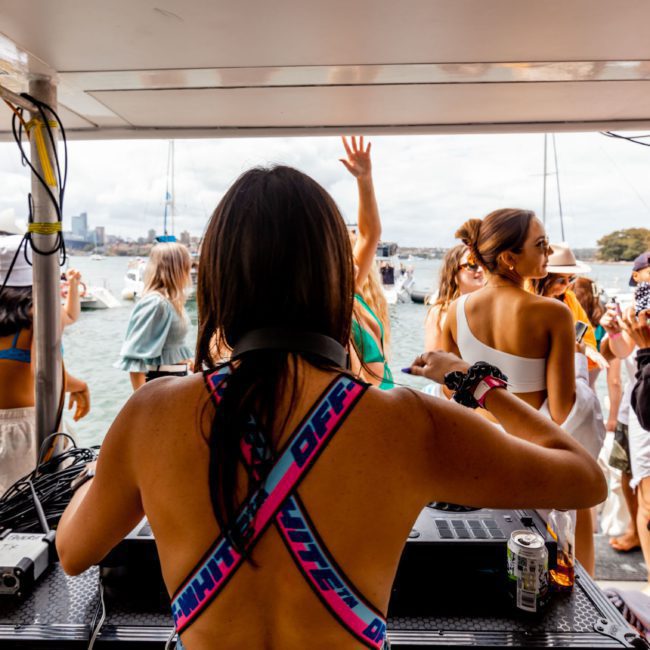 A DJ performing on a private yacht charter in Sydney Harbour with a crowd of people dancing and socializing on a sunny day by the water, with a bridge and city skyline visible in the background.