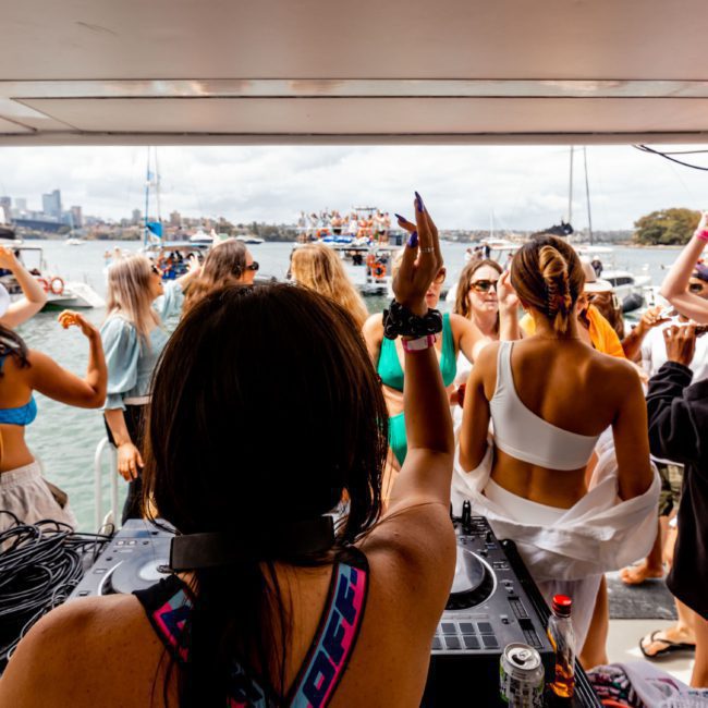 People dancing and enjoying music on a private yacht charter Sydney Harbour, with a DJ in the foreground and other boats in the background.
