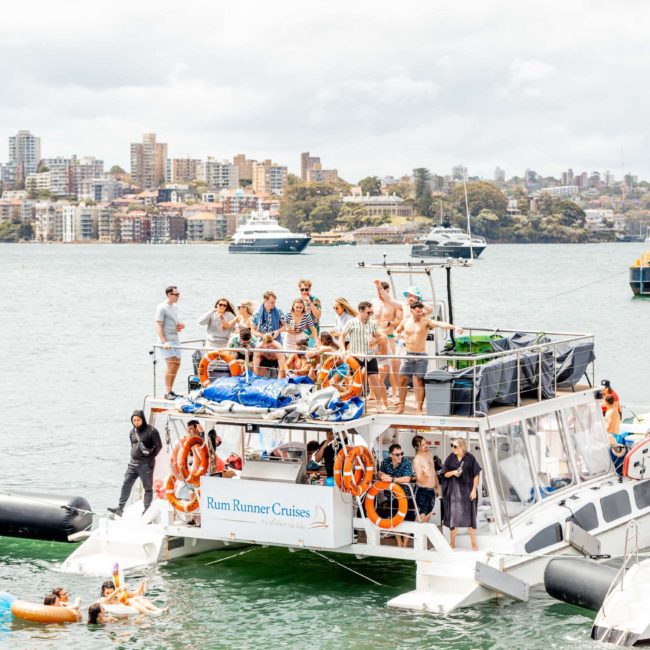 A group of people stand and sit on a large boat labeled "Rum Runner Cruises" docked near the coast, with buildings and a cloudy sky in the background. Other boats and people enjoying the water are visible, suggesting it could be a lively Catamaran party Sydney or part of corporate boat events Sydney.