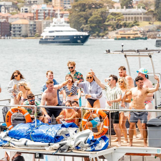 A group of people on a boat enjoys a sunny day on the water, with a cityscape and other boats in the background. The scene feels even more exhilarating with the Catamaran party Sydney experience adding to the joy.