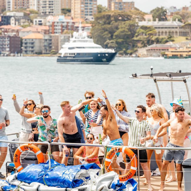 A group of people dancing and enjoying themselves on a private yacht charter in Sydney Harbour, with a background of buildings and other boats.