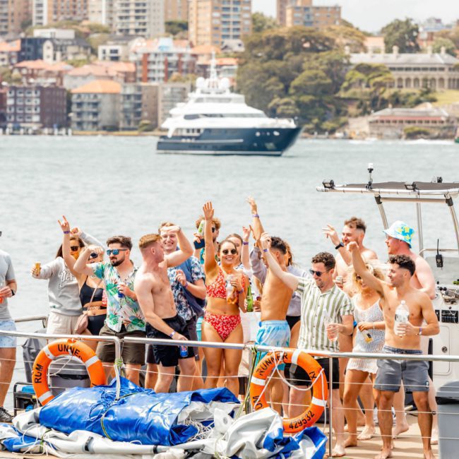 A group of people is celebrating on a catamaran with life rings and equipment. The background shows a cityscape and another boat on the water, perfect for a Sydney boat party hire.