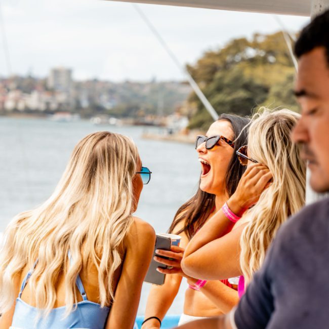 A man stands in the foreground, while three women wearing swimsuits and sunglasses converse and laugh on a luxury yacht hire Sydney, with a stunning cityscape and water in the background.