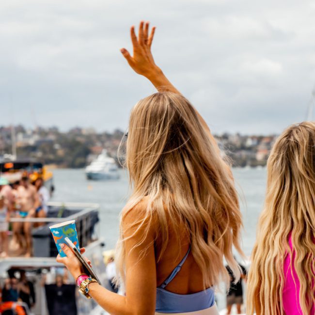 Two women with long blonde hair stand on a dock by the water, one waving towards a crowded boat in the background filled with people enjoying a Sydney boat party hire. A cityscape and cloudy sky are visible in the distance.