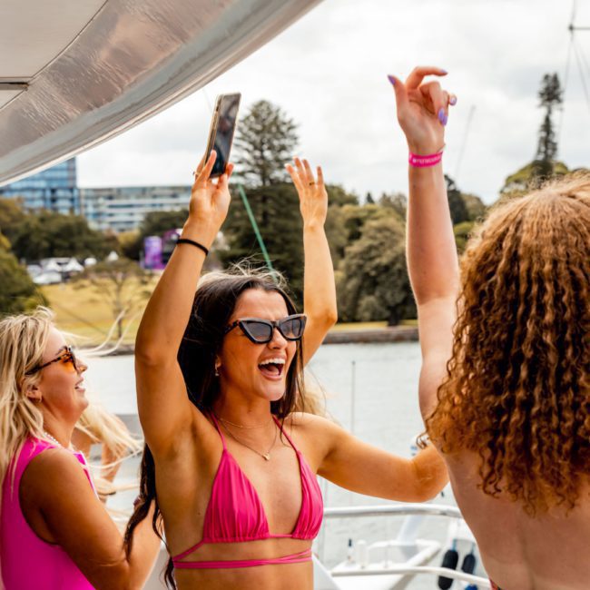 A group of people in swimwear is enjoying a lively boat party, with a woman in pink raising her hands, smiling and holding a phone, while others around her are cheerful. Trees and buildings are visible in the background. Experience the ultimate fun with Sydney boat party hire for your next event.